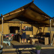 A woman stands on the deck of a safari tent with wooden furniture and laundry drying under clear skies.