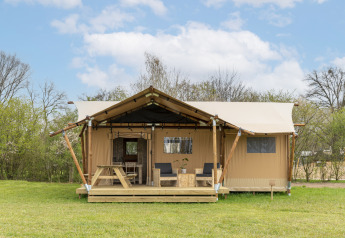 Foto van een safaritent met houten veranda, zitgedeelte en picknicktafel op een grasveld.
