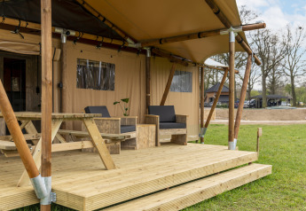 Tenda safari con veranda in legno, tavolo da picnic e sedie da salotto in un'area verde all'aperto.