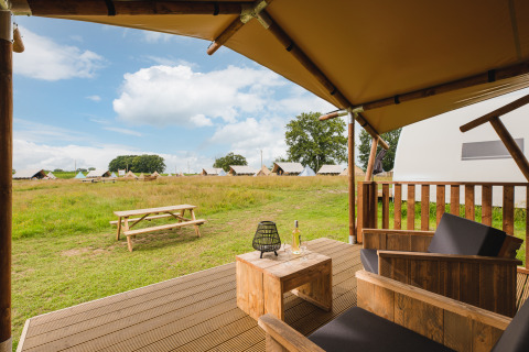 Vista desde una terraza de tienda safari con sillas y mesa de madera, vino y tiendas al fondo en un campo.