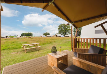 Vista desde una terraza de tienda safari con sillas y mesa de madera, vino y tiendas al fondo en un campo.