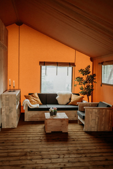 Accogliente salotto in tenda safari con mobili in legno, pareti arancioni, finestra e pianta decorativa.
