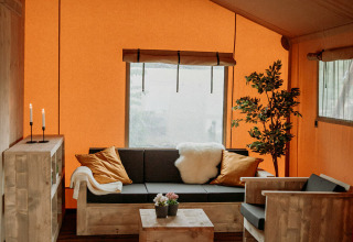Salon confortable dans une tente safari, avec meubles en bois, murs orange, grande fenêtre et plante décorative.