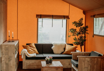 Knusse zithoek in een safaritent met houten meubels, oranje wanden, groot raam en decoratieve plant.
