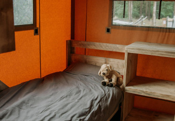 Das Innere eines Safari-Zeltes mit Einzelbett, grauer Bettwäsche, Stofftier und Holzmöbeln.
