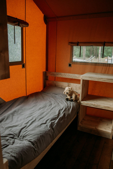 Binnenkant van een safaritent met een enkel bed, grijs beddengoed, knuffel en houten meubels.