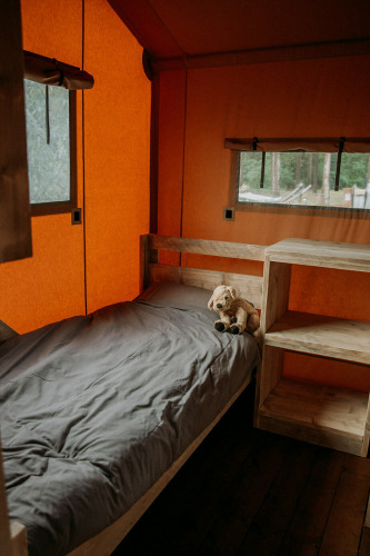 Interior de una tienda safari con cama individual, ropa de cama gris, peluche y muebles de madera.