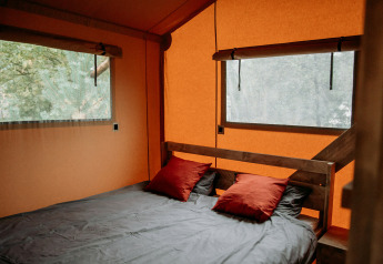 Interieur van een safaritent met een comfortabel bed, twee rode kussens en grote ramen met uitzicht.