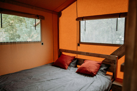 Interior of a safari tent featuring a cozy bed, two red pillows, and large windows with a view outside.
