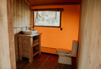Ein Badezimmer in einem Safarizelt mit Holzwänden, Toilette, Waschbecken und großem Fenster.