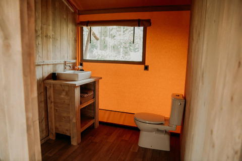 Baño dentro de una tienda safari, con paredes de madera, inodoro, lavabo y una gran ventana naranja.