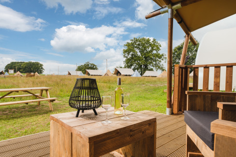 Hyggelig udsigt fra safari-teltterrasse med vin, glas og lanterne, picnicbord og telte i baggrunden.