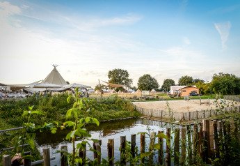 Glampingplek met tipitenten, zandstrand en vijver, omringd door groen onder een heldere blauwe lucht.