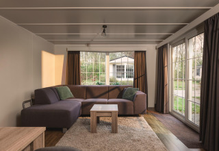 Lys opholdsstue i en bungalow med stor sofa, store vinduer og udsigt over ferieparken i Nederlandene.