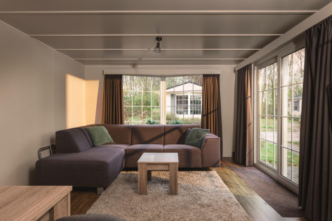 Helles Wohnzimmer in einem Bungalow mit großer Couch, großen Fenstern und Blick ins Grüne im Ferienpark.