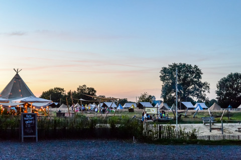 Glamping site at dusk with cozy tents, communal areas lit by string lights, and guests relaxing outdoors.