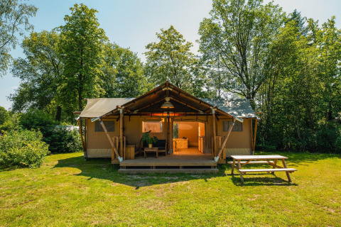 Foto van een grote safaritent met overdekt terras, omgeven door bomen en een picknickbank aan de voorkant.