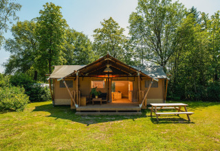 Foto van een grote safaritent met overdekt terras, omgeven door bomen en een picknickbank aan de voorkant.