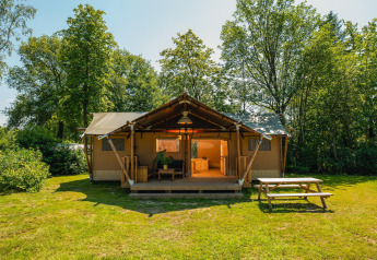 Foto af et rummeligt safari-telt med overdækket terrasse, omgivet af grønne træer og picnicbord foran.