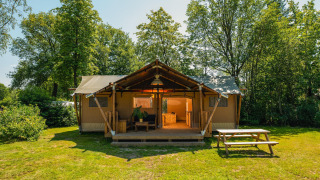 Foto af et rummeligt safari-telt med overdækket terrasse, omgivet af grønne træer og picnicbord foran.