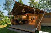 Outdoor view of a safari tent with wooden porch, seating area, and dining space, surrounded by greenery.