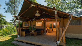 Vista exterior de una tienda safari con porche de madera, zona de estar y comedor, rodeada de naturaleza.