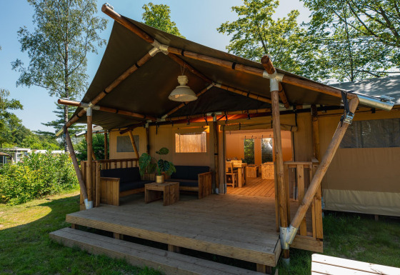 Vista exterior de una tienda safari con porche de madera, zona de estar y comedor, rodeada de naturaleza.