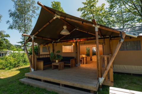 Vue extérieure d’une tente safari avec terrasse en bois, salon et salle à manger, entourée de verdure.