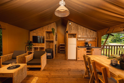 Binnenkant van safaritent met houten meubels, compacte keuken, salonhoek, tafel en buitenzicht.
