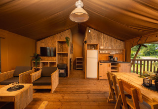 Binnenkant van safaritent met houten meubels, compacte keuken, salonhoek, tafel en buitenzicht.