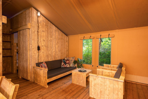 Intérieur d'une tente safari avec des meubles en bois rustiques et de grandes fenêtres sur la nature.