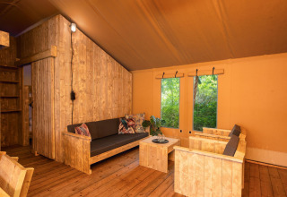 Safari tent interior featuring rustic wooden furniture, lounge area and large windows with forest views.