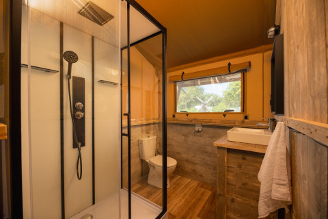 Baño en tienda safari con ducha acristalada, inodoro, lavabo de madera y ventana con vistas naturales.