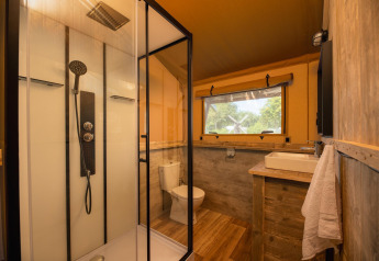 Bagno in tenda safari con ampia doccia in vetro, wc, lavabo in legno e una finestra panoramica sul verde.
