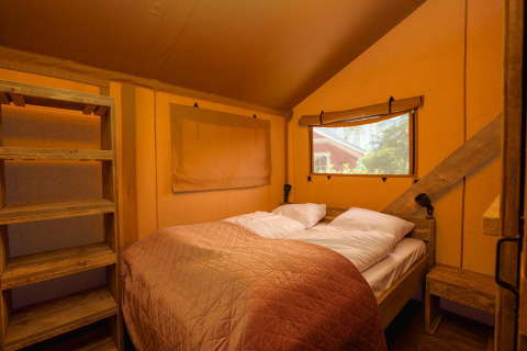 Interior de tienda safari con cama doble, estantería de madera, ventana y luz cálida en el dormitorio.