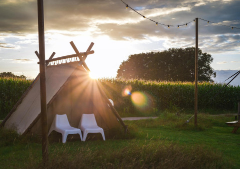 Tente de glamping au coucher du soleil avec deux chaises blanches, entourée de champs et guirlandes lumineuses.