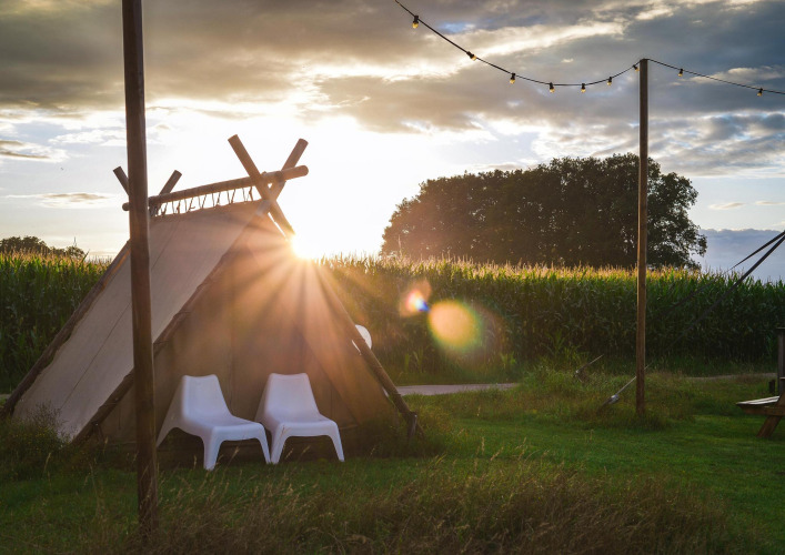 Tienda de glamping al atardecer con dos sillas blancas frente a un campo y luces colgantes encima.