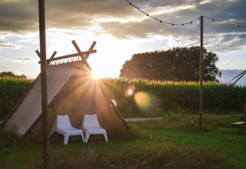 Tente de glamping au coucher du soleil avec deux chaises blanches, entourée de champs et guirlandes lumineuses.