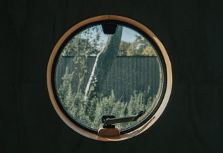 Ventana redonda en Wikkel tiny house de De Klepperstee, Países Bajos, con vista al jardín y arbustos.