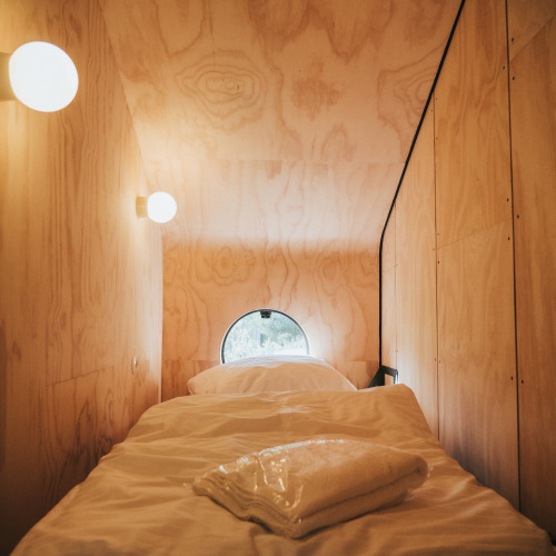 Alojamiento de glamping con cama individual, paredes de madera, ventana redonda y luces suaves.