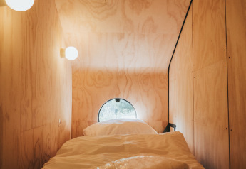 Alojamiento de glamping con cama individual, paredes de madera, ventana redonda y luces suaves.
