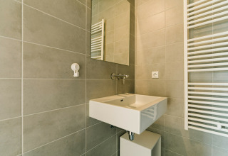 Baño moderno con azulejos grises, lavabo blanco, espejo y radiador toallero en una casa natural.