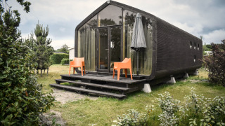 Tiny house noire moderne avec façade vitrée, chaises orange et parasol, dans la nature à De Klepperstee.