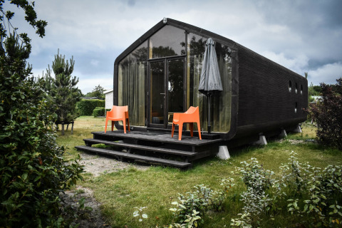 Modern black tiny house with glass front, orange chairs and umbrella, surrounded by nature at De Klepperstee.