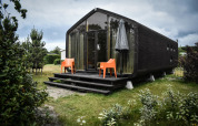 Casa pequeña negra moderna con fachada de vidrio, sillas naranjas y sombrilla en la naturaleza de De Klepperstee.