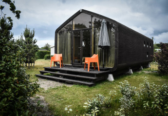 Casa pequeña negra moderna con fachada de vidrio, sillas naranjas y sombrilla en la naturaleza de De Klepperstee.
