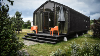 Casa pequeña negra moderna con fachada de vidrio, sillas naranjas y sombrilla en la naturaleza de De Klepperstee.