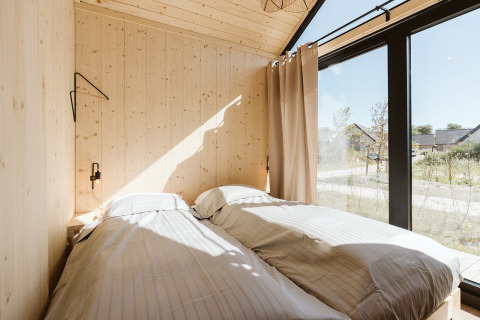 Dormitorio soleado en Stulp, tiny house en De Klepperstee, con grandes ventanales y paredes de madera clara.