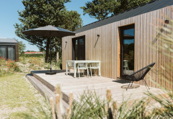Udendørs træterrasse ved et lille hus på De Klepperstee, med havemøbler og parasol under blå himmel.