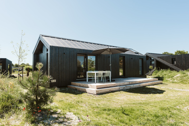 Casa pequeña negra con terraza de madera y muebles de exterior en De Klepperstee, Países Bajos, día soleado.